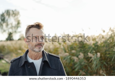 Serious man standing thinking outdoors at the coast alongside tall reeds looking off to the side with a contemplative expression