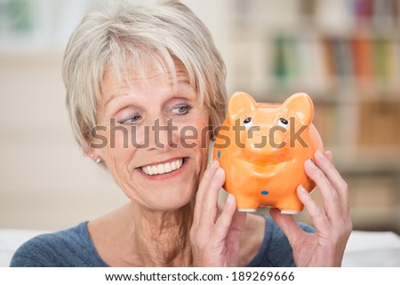 Charismatic elderly woman holding up a piggy bank looking sideways at is as she smiles while contemplating what to do with her savings