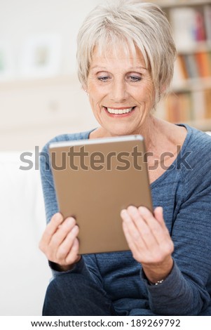 Attractive senior woman enjoying an e-book smiling as she reads the story while relaxing at home in her living room