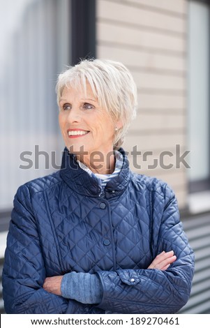 Confident friendly stylish senior woman standing wearing a high necked jacket with folded arms outside her house looking to the side with a smile