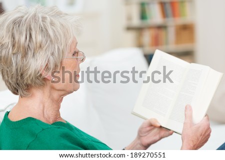 Senior woman relaxing reading a book on a sofa at home, over the shoulder view