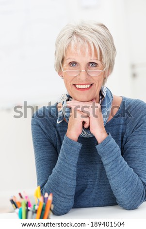 Beautiful friendly senior woman sitting at a desk in the office wearing her spectacles and a trendy scarf smiling at the camera