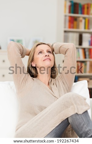 Pretty middle-aged woman sitting daydreaming on a sofa in her living room with her head tilted back and hands clasped behind her head