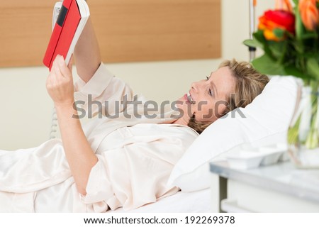 Smiling happy woman patient reading a book in hospital as she relaxes comfortably on the pillows in the hospital bed