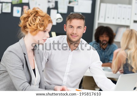 Smiling confident young businessman sitting sharing a desk with an attractive redhead woman as they work together on a laptop computer