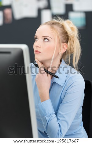 Young businesswoman sitting thinking at her desk with her hand holding a pen to her chin and her eyes raised in contemplation