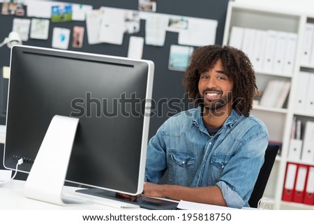 Charismatic African American businessman with an afro hairdo sitting at his desk with a desktop monitor smiling at the camera