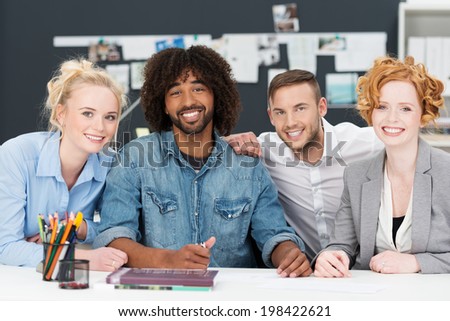 Happy smiling casual young African American businessman with his team of young men and women sitting in a row at a desk smiling at the camera