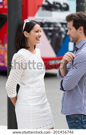 Stylish young couple standing chatting in the street with the woman laughing as she leans back against a pole in a fresh white summer dress