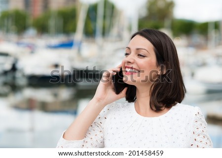 Beautiful woman smiling as she chats on her mobile phone looking away into the distance with a beaming smile of pleasure, marine harbour in the background