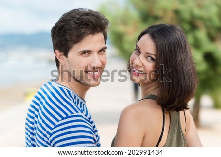Attractive young couple on a tropical beach turning back to look at the camera over their shoulders with happy warm smiles