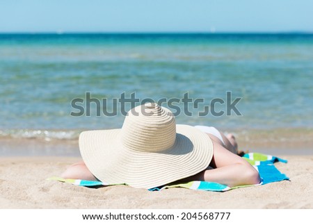 Woman sunbathing on a hot tropical summer beach lying on her towel on the sand hidden under her wide brimmed straw sunhat against an ocean backdrop