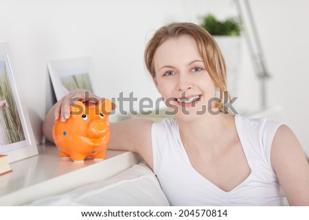 Beautiful natural smiling young woman holding onto a piggy bank as she relaxes on a couch at home showing her awareness of the importance of saving