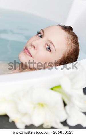 Beautiful young woman relaxing in her bath with white flowers in the foreground looking over the top of the curved rim with a peaceful look of wellbeing