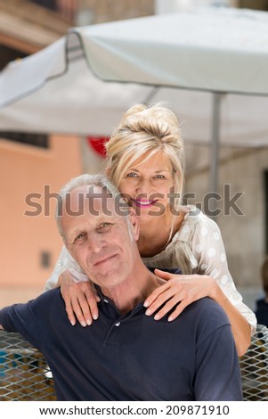 Happy attractive middle-aged couple relaxing outdoors on a patio under an umbrella with the wife leaning over her husbands shoulders from behind to smile at the camera