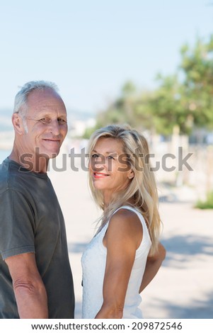 Middle-aged couple on vacation at the coast smiling as they turn to look at the camera while walking on a seafront promenade