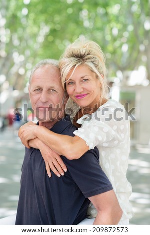 Middle-aged man giving an attractive smiling woman a piggy back ride as she hugs him around the shoulders on a hot summer day in town