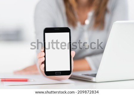 Businesswoman displaying a blank mobile phone with a white screen towards the camera as she sits at her desk, closeup of her hand with focus to the phone