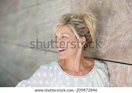 Vivacious beautiful blond woman posing leaning against a receding wall as she looks to the left with a laughing smile