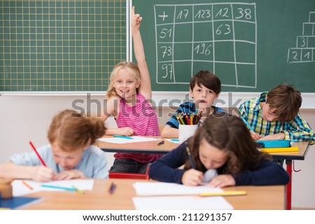 Intelligent enthusiastic little girl in class holding up her hand with a happy smile to attract attention and answer a question