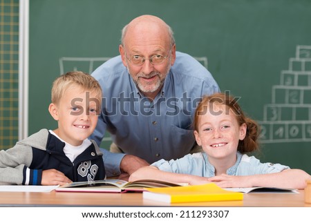 Two happy young children with their elderly male teacher in the classroom posing together in front of the blackboard smiling at the camera