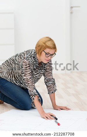 Young woman working on a floor plan in her new house or apartment kneeling on the floor working over a blueprint with marker pens