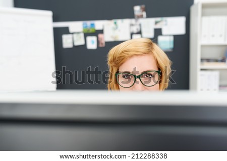 Businesswoman wearing heavy rimmed glasses looking over the top of her computer monitor at the camera