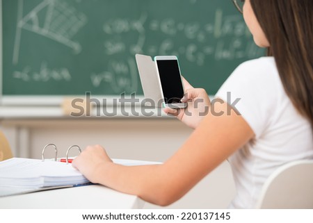 Over the shoulder view of a young female student using a mobile phone in class with the blank screen visible to the viewer