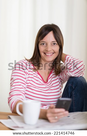 Smiling housewife relaxing over morning coffee sitting at the dining table with her mobile phone and the newspaper