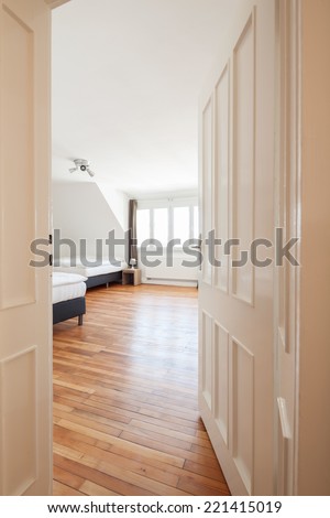 View through the door past white paneling into a spacious airy loft bedroom with two double beds, windows and a wooden parquet floor