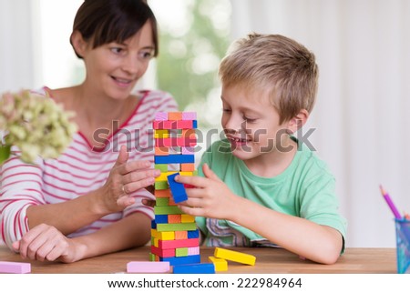 Young mother helping her small son build a tower from colorful building blocks as they sit together at the dining table at home