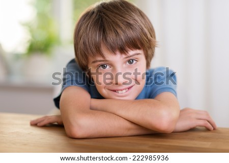 Handsome little boy with a lovely wide smile leaning his head on his arms on a wooden table smiling at the camera
