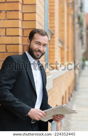 Confident bearded businessman relaxing with a newspaper during his lunch break leaning against the exterior wall of an old brick office building