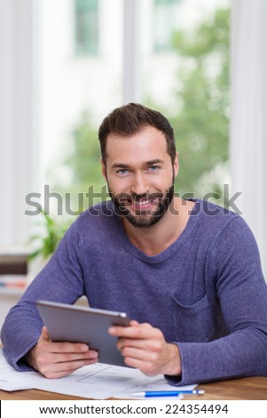 Smiling happy man working with a handheld tablet computer at his desk in front of a window