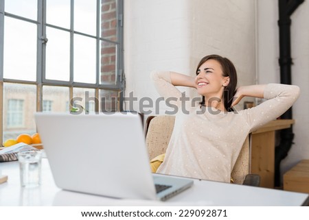Happy woman relaxing in her kitchen with her laptop on the counter looking out of a window with a lovely smile