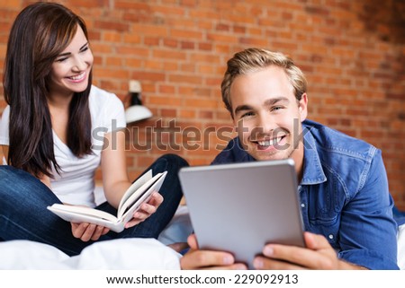 Happy Young White Couple Holding Book and Tablet Gadget While on the Bed.