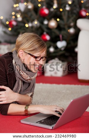 Woman wearing glasses with a notebook computer lying in front of a Christmas tree smiling as she surfs the internet and social media