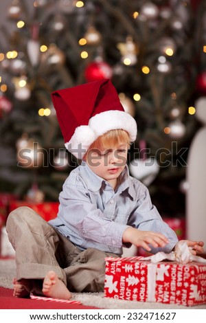 Cute little redhead boy in a Santa hat sitting on the floor in front of the decorated tree opening a Christmas gift