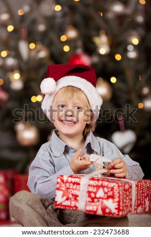 Happy adorable small boy opening an Xmas gift sitting in front of the Christmas tree with a gift-wrapped box on his lap smiling at the camera