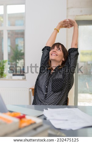 Contented successful businesswoman sitting at her desk in the office stretching her hands above her head with a smile of satisfaction