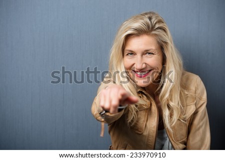 Vivacious attractive blond middle-aged woman pointing at the camera with a beaming playful smile, blackboard background with copyspace