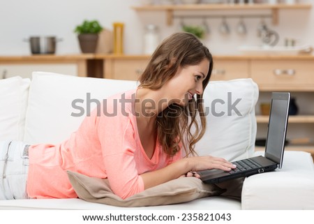 Young woman relaxing at home surfing the internet on her laptop computer as she lies on a comfortable sofa