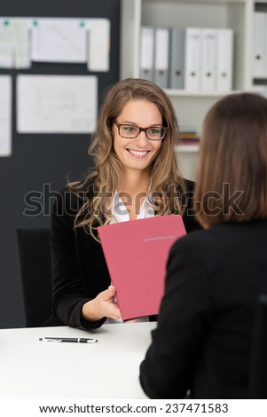 Happy Businesswoman with Long Blond Hair Giving Reports in Dark Pink Folder to her Co-worker at her Worktable.