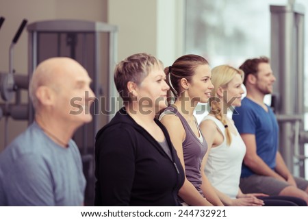 Row of diverse young and old people in a gym sitting listening to the instructor with focus to an elderly lady in the centre in a healthy lifestyle concept