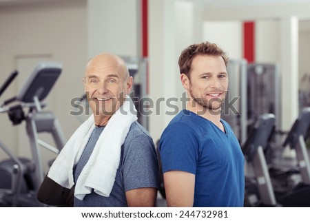 Two fit healthy men posing back to back at the gym, one senior and one young, looking at the camera with smiles full of vitality in a health and fitness concept