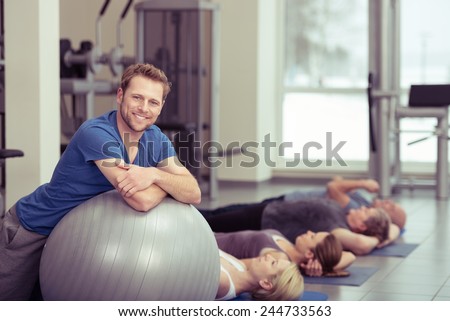 Smiling Fit Young Man Leaning on Gray Large Exercise Ball with Women at the Back Exercising. Captured Inside the Fitness Gym.