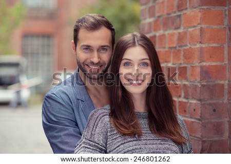 Happy attractive couple in an urban street standing in a close embrace facing the camera with warm friendly smiles against a brick wall