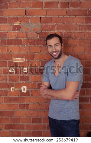 Confident young bearded man standing with folded arms leaning on a red brick wall smiling at the camera, with copyspace