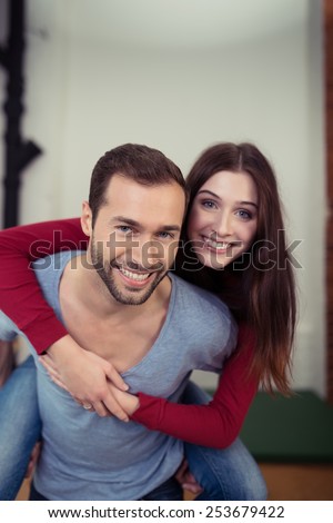 Playful young couple romping at home with the young man giving his wife a piggy back ride as they laugh at the camera