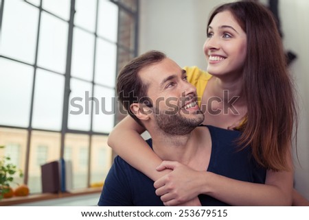 Happy loving young couple sitting at home with the young woman hugging her husband from behind as she looks out of the window with a smile, watched by him